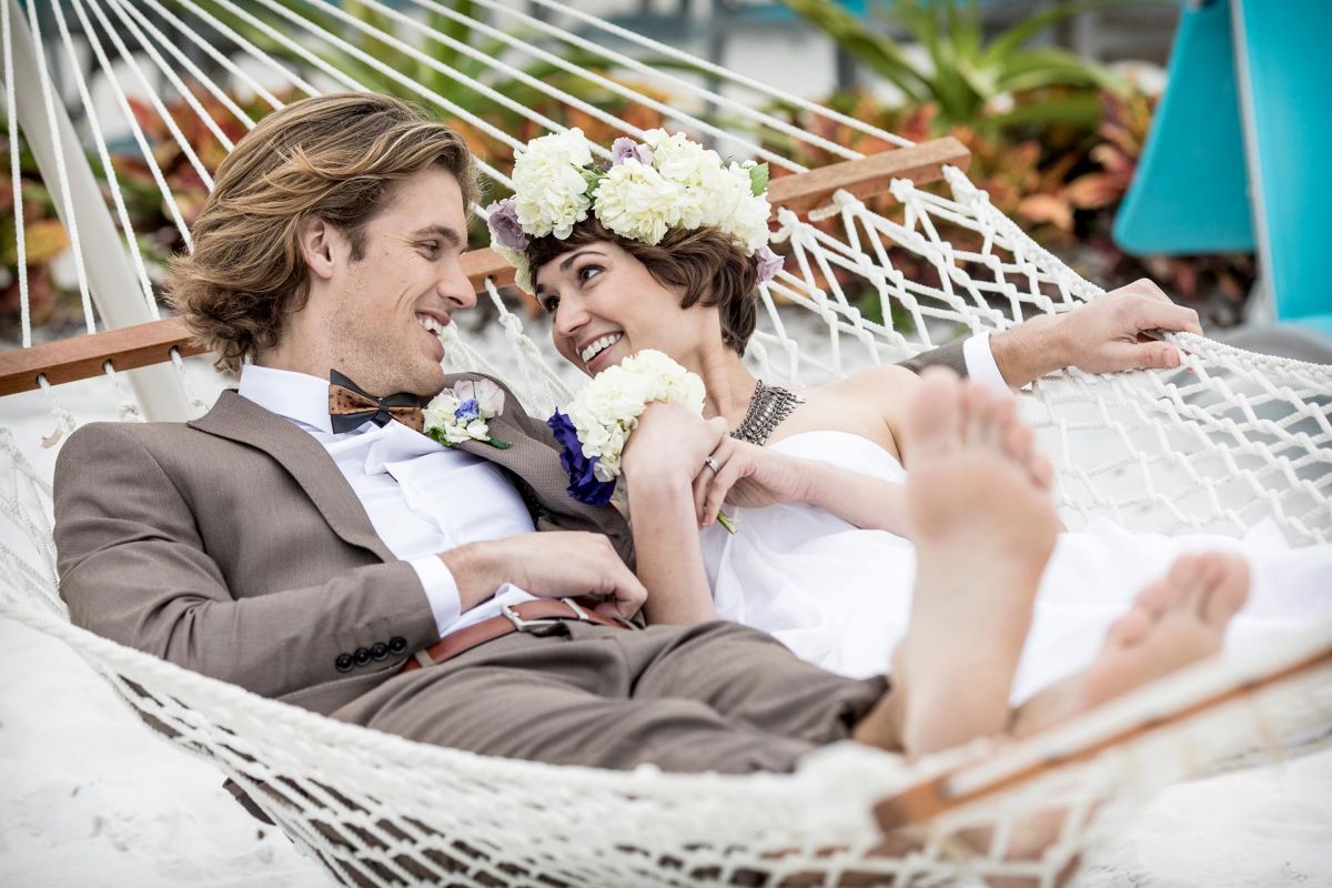 Bride and groom lying on a hammock on the sand in their wedding outfits.