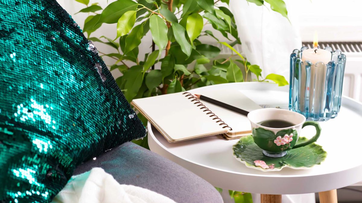 Notepad and pencil on a table with a cup of tea and a sequin throw pillow on a chair.