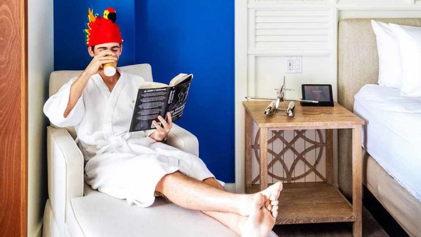Man lounging on a comfy chair and reading a book while wearing a parrot hat