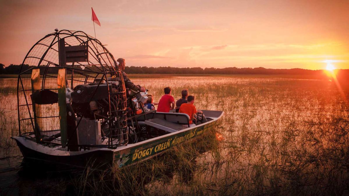 People riding an airboat on the marshy waters of Boggy Creek at sunset.