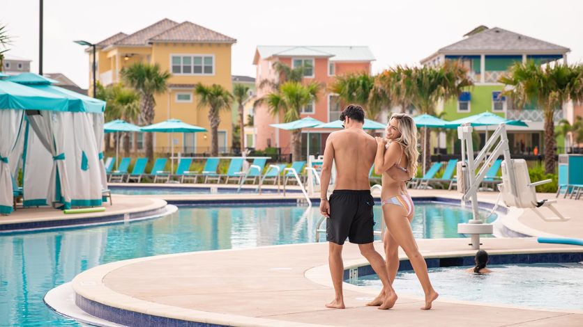 Man and woman in swim suits walking along the Margaritaville Resort Orlando pool, with the colorful Margaritaville Cottages in the background.