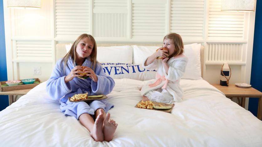 Two young girls eating burgers and fries on a bed in their Margaritaville Resort Orlando hotel room.
